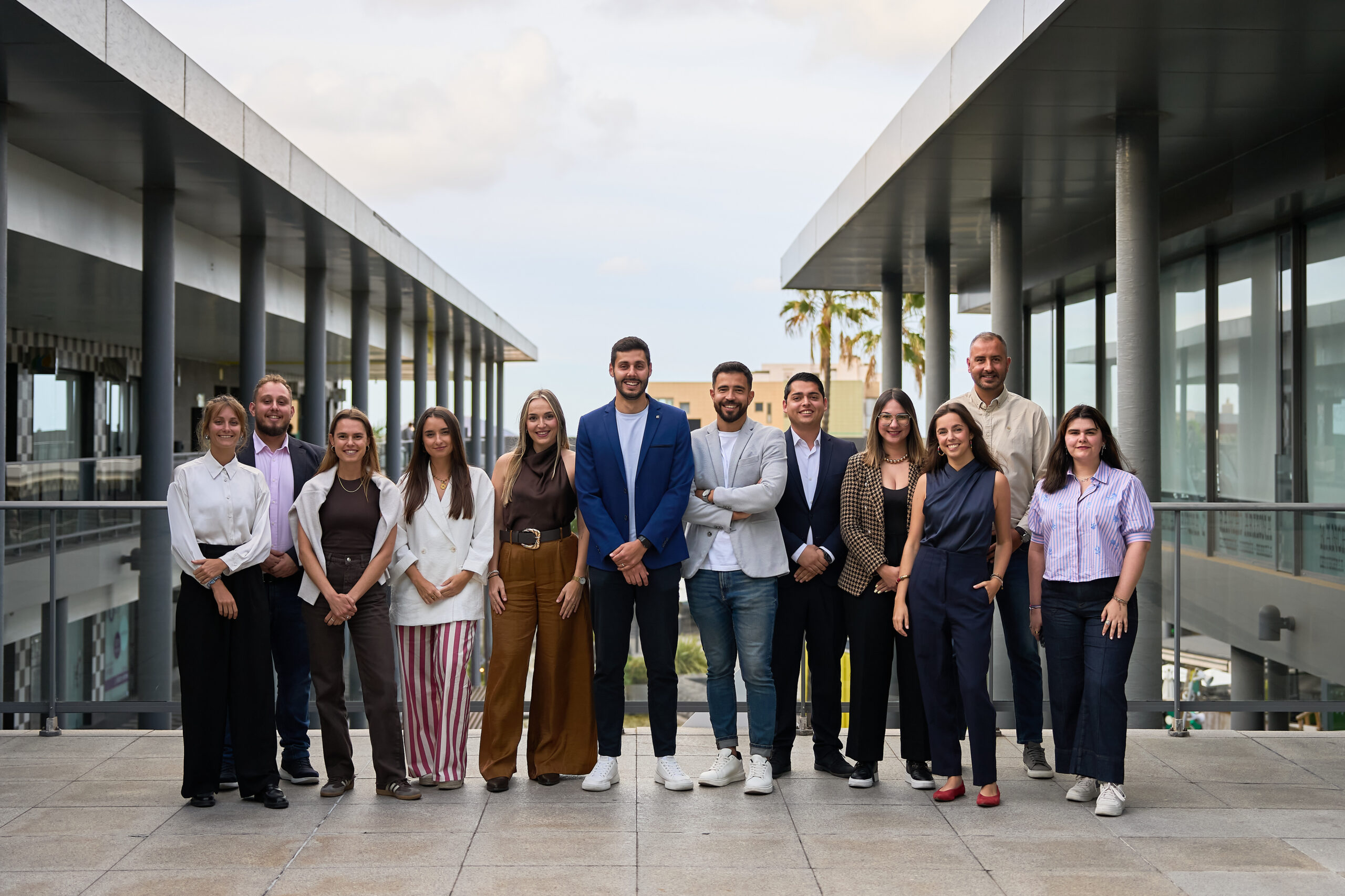 Grupo de profesionales posando en un entorno moderno, con arquitectura contemporánea y palmeras al fondo. Todos están vestidos de manera formal y sonríen, mostrando un ambiente colaborativo y amigable.