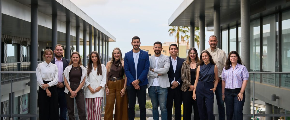 Grupo de profesionales posando en un entorno moderno, con arquitectura contemporánea y palmeras al fondo. Todos están vestidos de manera formal y sonríen, mostrando un ambiente colaborativo y amigable.